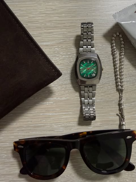 Steel watch with a green dial, tortoiseshell Wayfarer sunglasses, and a leather wallet
